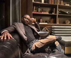 Eleazar Adjehoun sits on a leather couch in a lounge setting, dressed in a blazer and glasses, resting his arm on the chair.