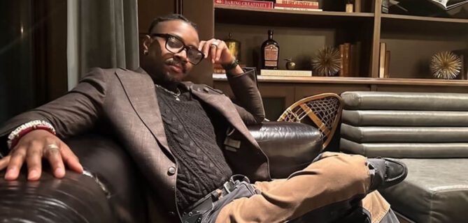 Eleazar Adjehoun sits on a leather couch in a lounge setting, dressed in a blazer and glasses, resting his arm on the chair.