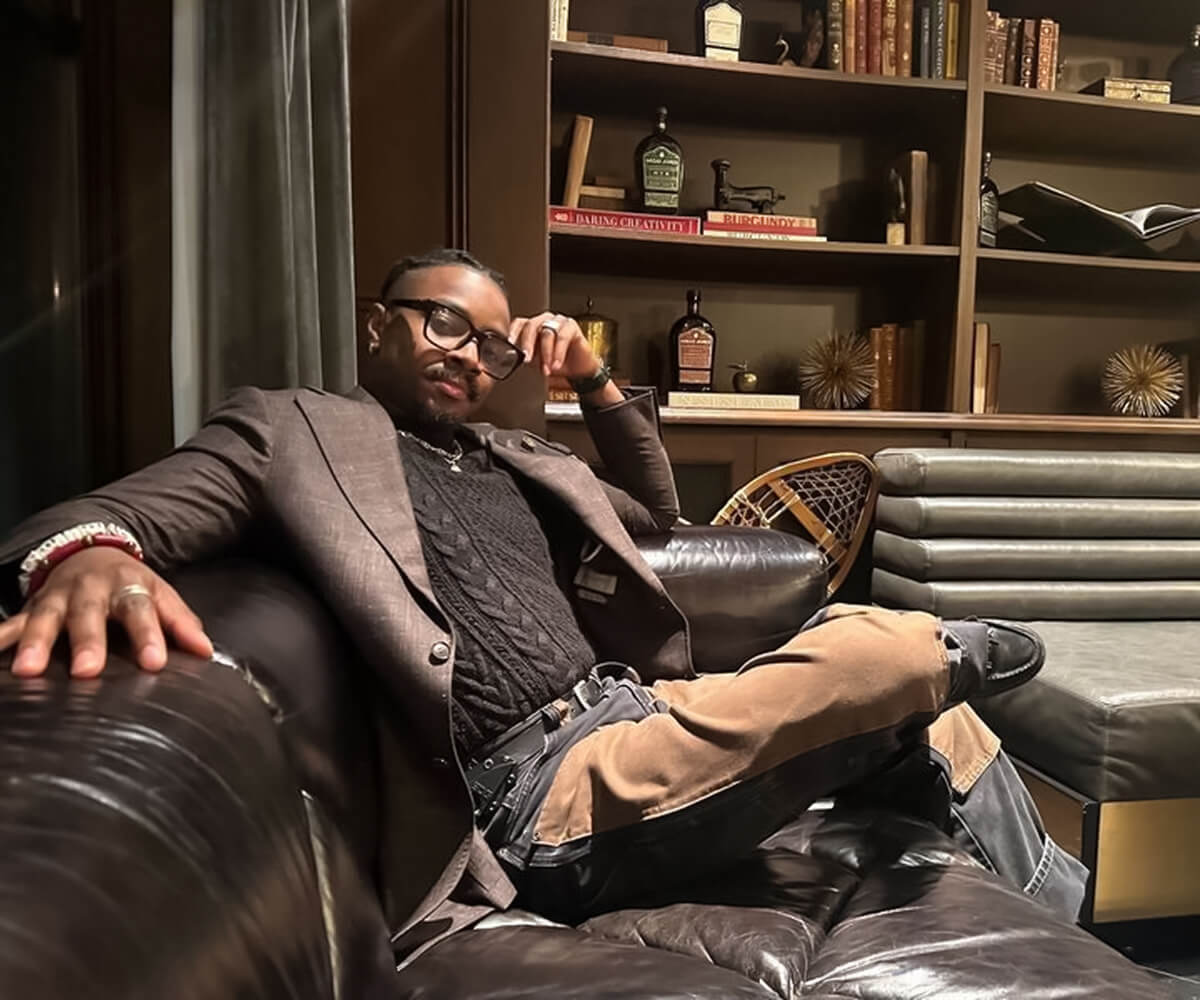Eleazar Adjehoun sits on a leather couch in a lounge setting, dressed in a blazer and glasses, resting his arm on the chair.