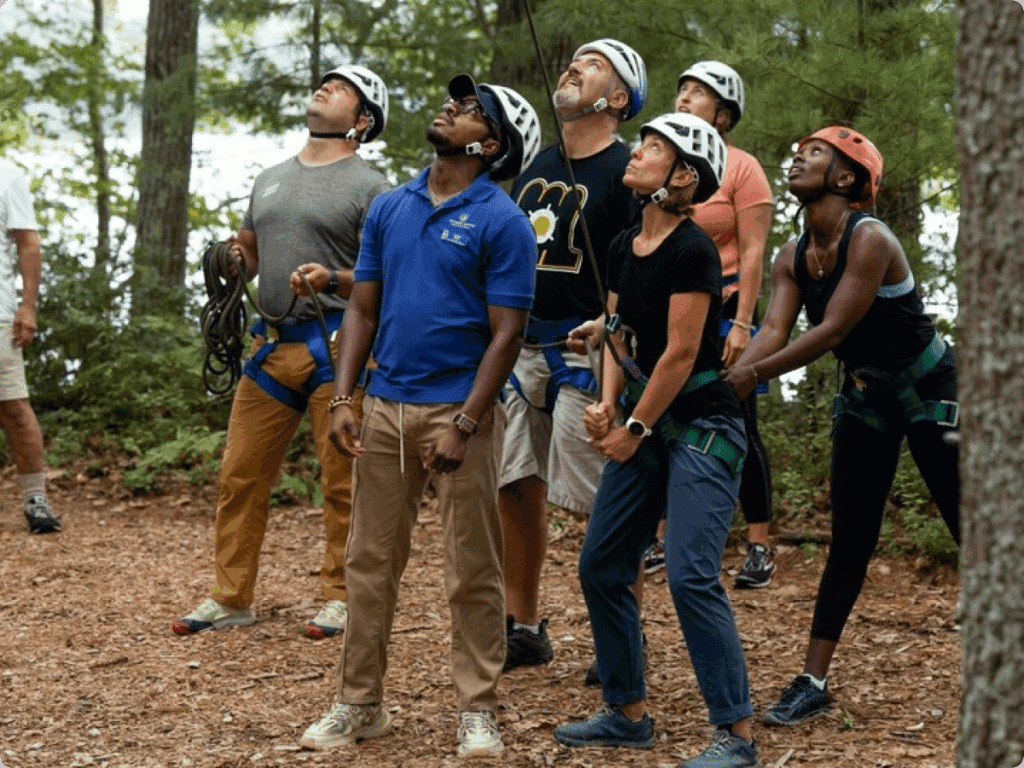 A diverse group of adults wearing helmets and harnesses look upward together during an outdoor ropes activity.