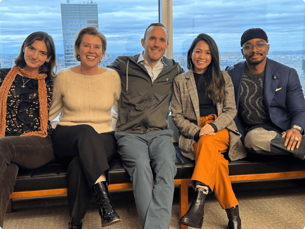 Eleazar Adjehoun sits with colleagues on a bench indoors, smiling together in a casual group photo.