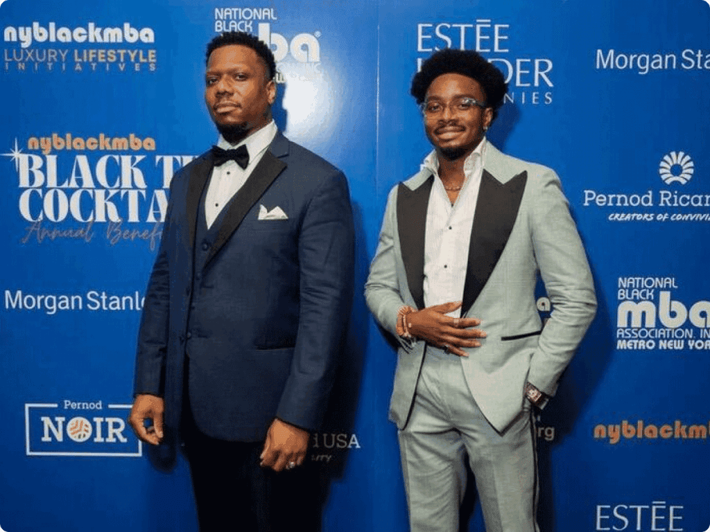 Eleazar Adjehoun stands at a formal event wearing a light-colored suit, posing for a photo with another attendee in front of a branded backdrop.