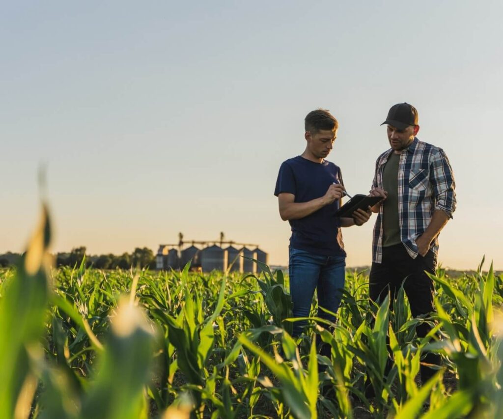 Two men standing in a green crop field at sunset, reviewing information on a tablet with farm buildings visible in the background.