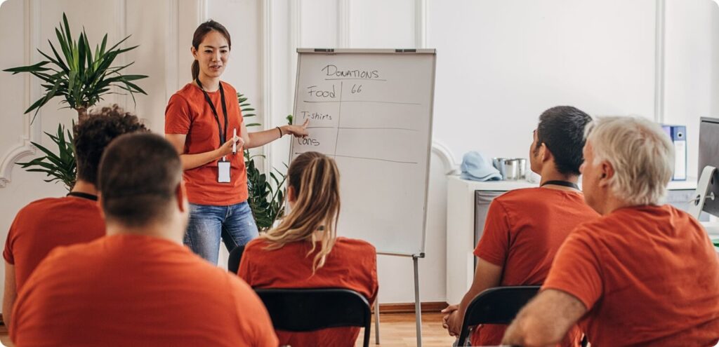 A volunteer facilitator points to a whiteboard listing donation items while meeting with a group of volunteers.
