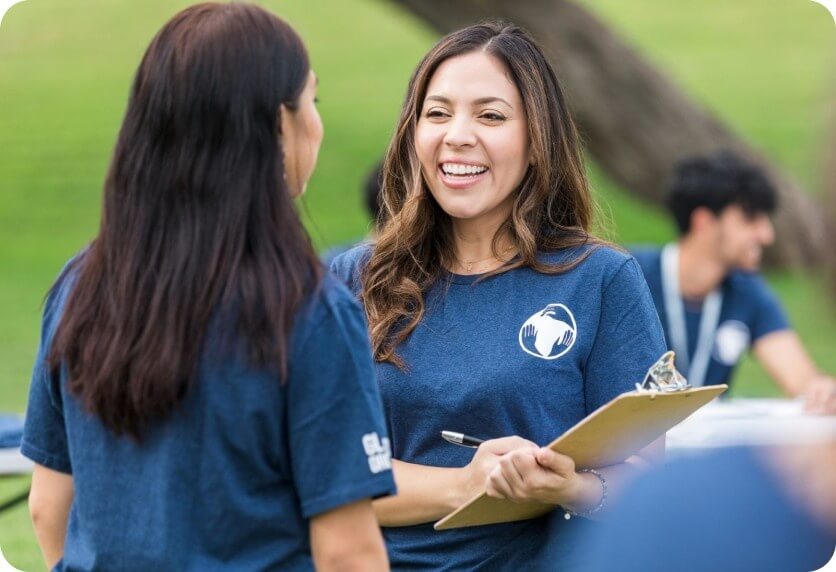 A volunteer coordinator smiles while reviewing notes on a clipboard and talking with another volunteer in a park setting.