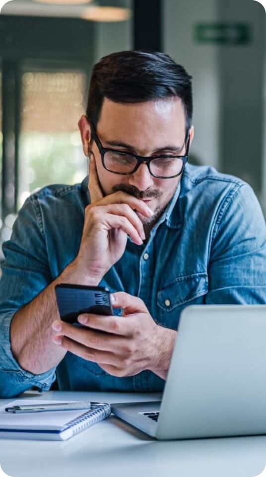 A person reviews information on a smartphone while working at a desk with a laptop and notebook.