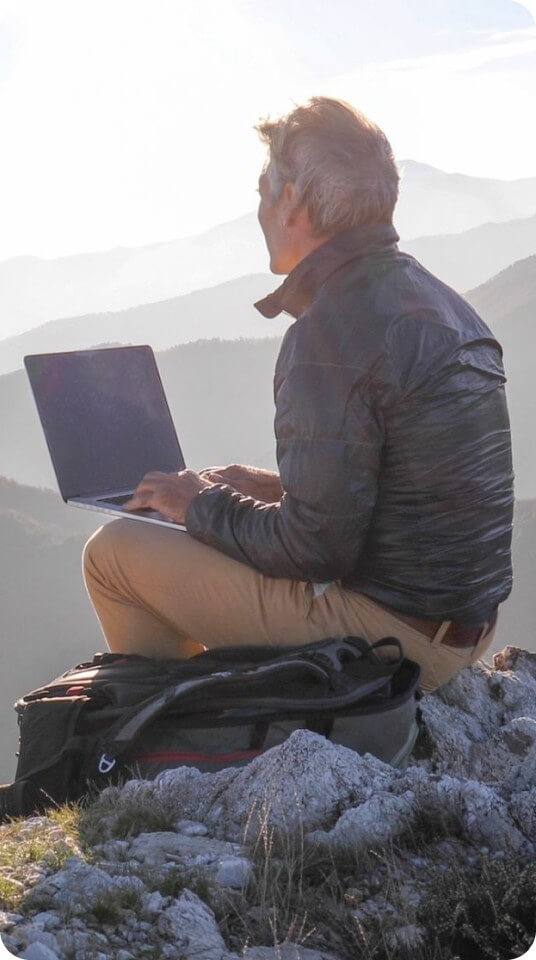 A person sits outdoors using a laptop with a mountain landscape in the background.