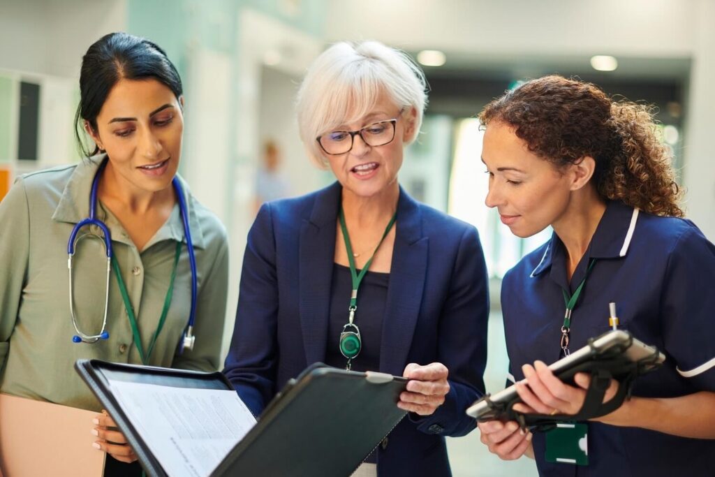 Three healthcare professionals in a hospital setting review a clipboard together, with one holding a tablet-style device and another wearing a stethoscope.