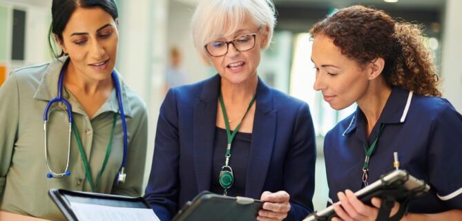 Three healthcare professionals in a hospital setting review a clipboard together, with one holding a tablet-style device and another wearing a stethoscope.