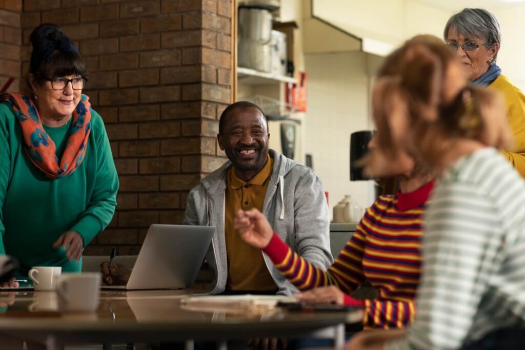 Alt text: Group of adults gathered around a table in a community space, smiling and talking while one person uses a laptop during a collaborative meeting.