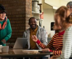 Alt text: Group of adults gathered around a table in a community space, smiling and talking while one person uses a laptop during a collaborative meeting.