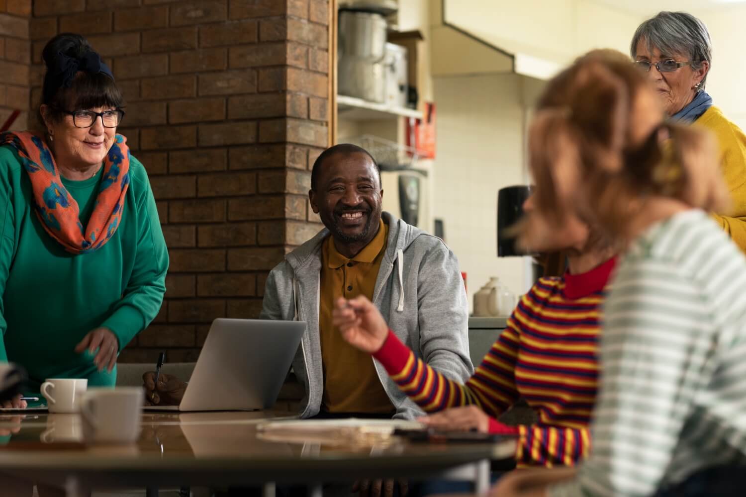 Alt text: Group of adults gathered around a table in a community space, smiling and talking while one person uses a laptop during a collaborative meeting.