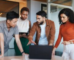 Four coworkers in a modern office gather around a laptop, discussing something on the screen while one person holds a tablet.