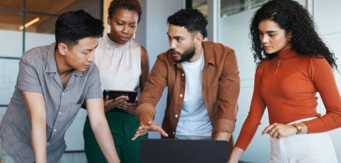 Four coworkers in a modern office gather around a laptop, discussing something on the screen while one person holds a tablet.