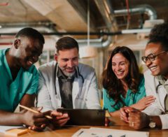 Four healthcare professionals in scrubs and lab coats gathered around a tablet, smiling and reviewing information together in a clinical office setting.
