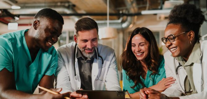 Four healthcare professionals in scrubs and lab coats gathered around a tablet, smiling and reviewing information together in a clinical office setting.