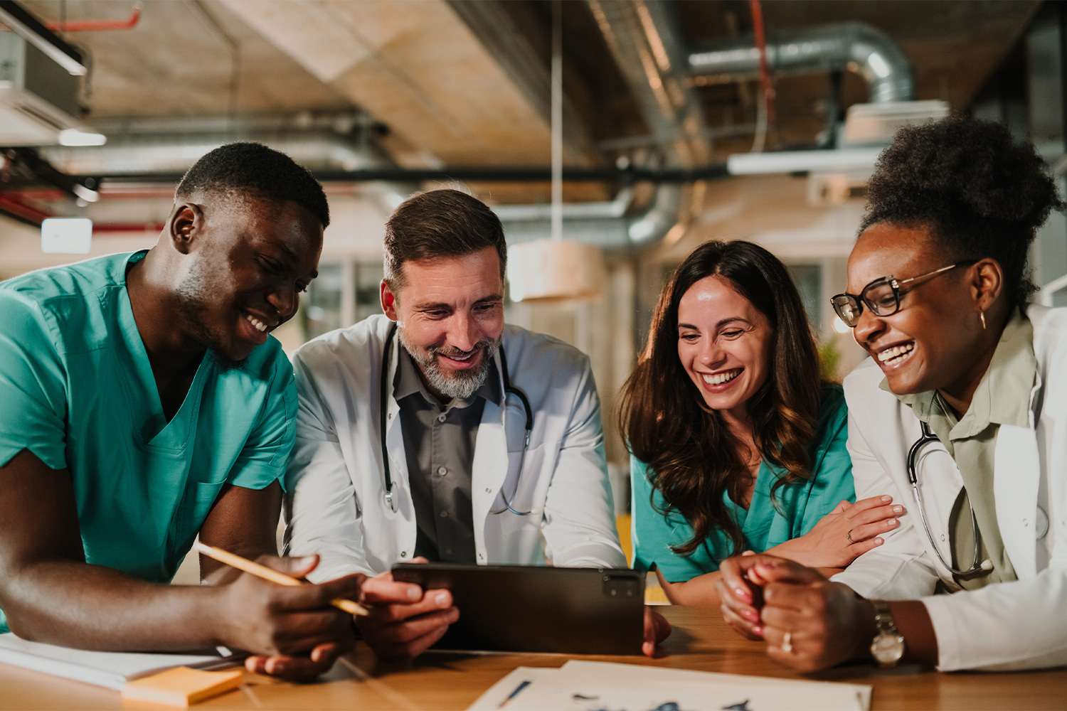 Four healthcare professionals in scrubs and lab coats gathered around a tablet, smiling and reviewing information together in a clinical office setting.