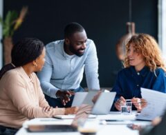 Three colleagues review documents and a laptop together while talking at a table in a modern office.