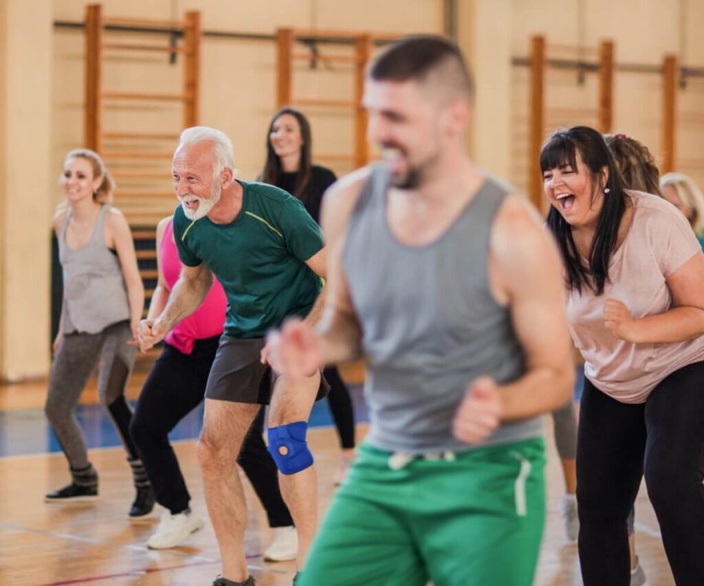A lively group fitness class in a gym, with participants smiling and moving through an exercise together.