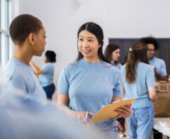 A volunteer holding a clipboard talks with another volunteer while others organize supplies in the background.