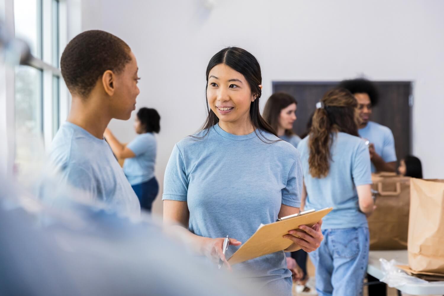 A volunteer holding a clipboard talks with another volunteer while others organize supplies in the background.