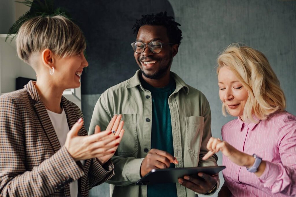 Three colleagues stand together smiling while looking at a tablet in an office setting.