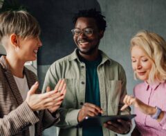 Three colleagues stand together smiling while looking at a tablet in an office setting.