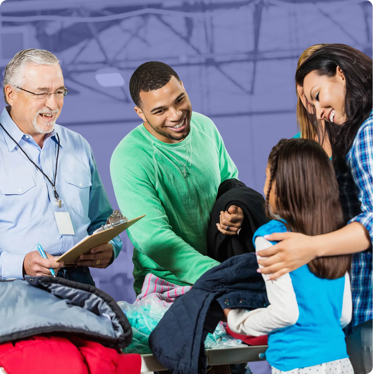 Group of volunteers smiling as they distribute coats to a child, with one person holding a clipboard and a family receiving donated clothing indoors.